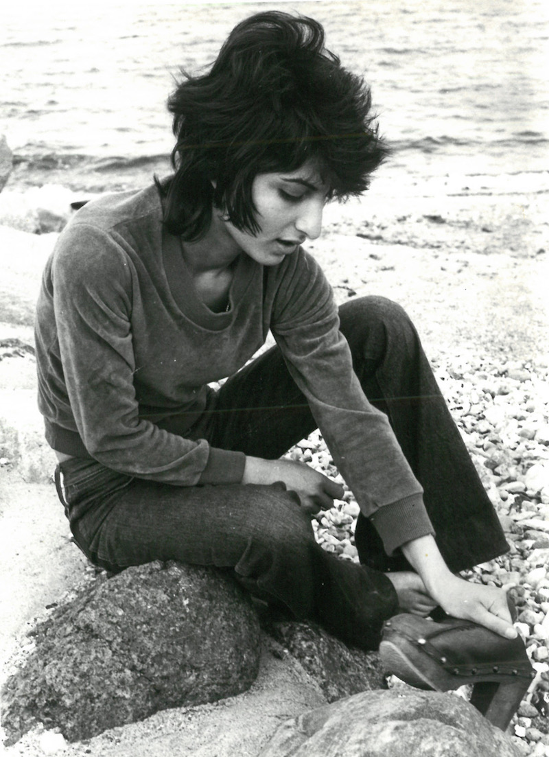 a black and white photograph of the poet Semra Ertan sitting on a log at the beach, looking down at a rock in her hand