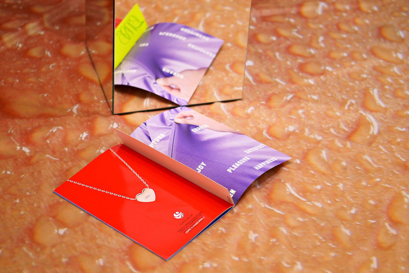 a close up of the open book with a heart shaped locket on a red page, the book is sitting next to a mirrored plinth and on a floor covering that looks like water droplets