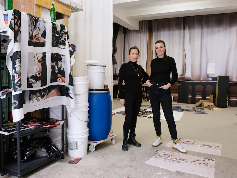 two artists stand in their studio space, next to shelves full of materials, both dressed in black