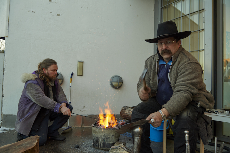 two people, one with a black cowboy hate, sit around a small fire in front of a government building