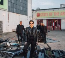 a woman in full black leather and sunglasses stands authoritatively in front of a lot full of car parts and scrap metal, with several family members behind her and a sign in the background reading "state of the arts"