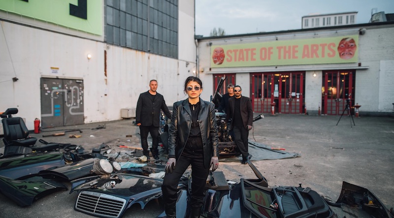 a woman in full black leather and sunglasses stands authoritatively in front of a lot full of car parts and scrap metal, with several family members behind her and a sign in the background reading "state of the arts"