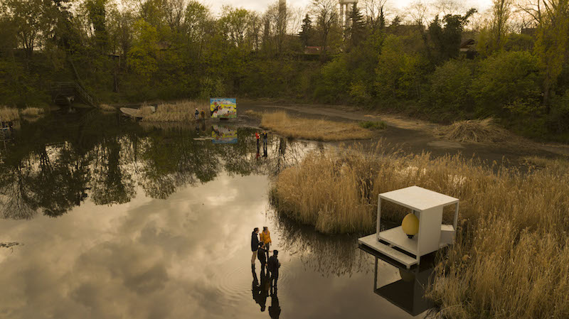 a drone aerial photo of the water basin, with art installations spread across the water and people in rain boots looking at them