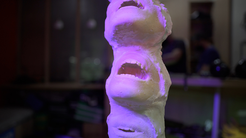 a close up shot of a tower sculpture made up of open mouths, and bathed in purple light