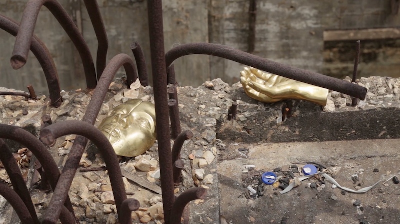 a construction site with metal sticking out of concrete and two gilded body parts lying on top, a hand and a face