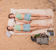 twin girls wearing matching blue shorts, sage green crop tops and long braids lie on a beach towel on the sand