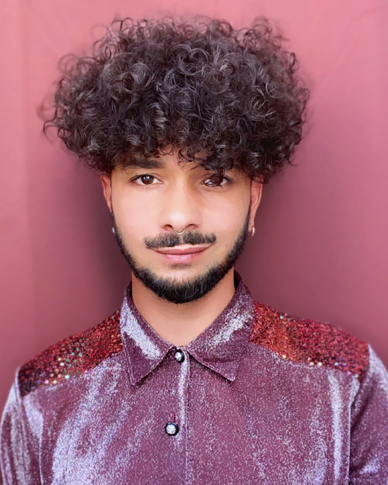 a person with curly dark hair and a dark beard wears a shiny purple shirt in front of a pink wall