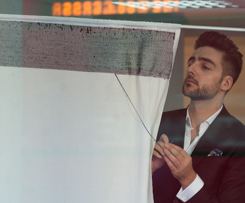 the artist stands behind a hanging piece of white fabric, embroidering black thread onto it from the side