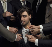 the artist sits in the middle of the photo's frame, staring into the camera, while several men's hands in suits reach out to light a cigarette in his mouth