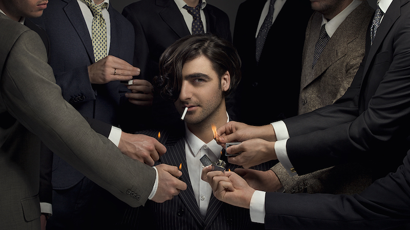 the artist sits in the middle of the photo's frame, staring into the camera, while several men's hands in suits reach out to light a cigarette in his mouth