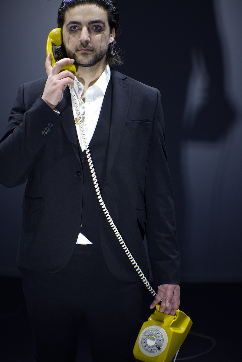 the artist stands in a room, visible from the window outside on the street, wearing a black suit and white shirt, with mascara running down his face and a yellow rotary telephone in his hand