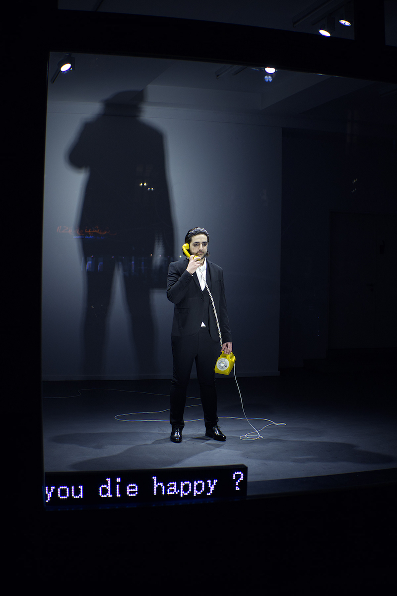 the artist stands in a room, visible from the window outside on the street, wearing a black suit and white shirt, with mascara running down his face and a yellow rotary telephone in his hand