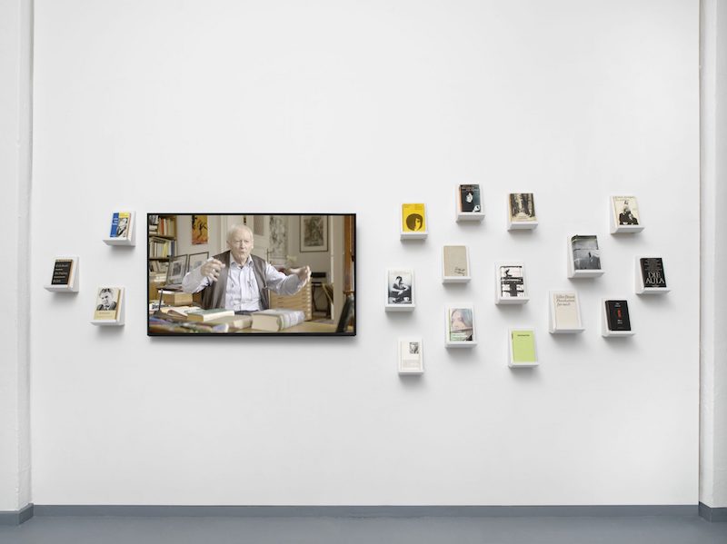 the white gallery wall with small shelves the size of individual books that they hold, and a screen showing a video interview of an older man at a desk