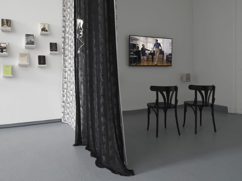 the gallery room divided with a long black curtain, on one side are books mounted on small shelves on the wall and on the other is a film screening on a small tv, with two chairs in front of it