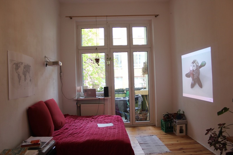 a red bed in an empty bedroom faces a projection on a wall, in front of a balcony door