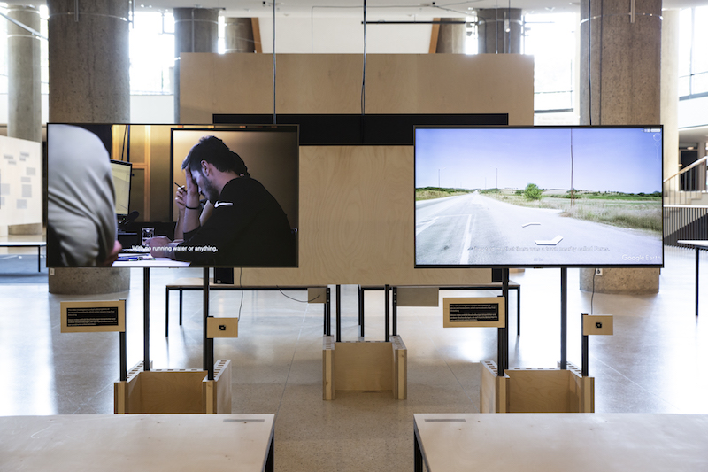a photo of an exhibition installed in HKW with several screens and desks with chairs