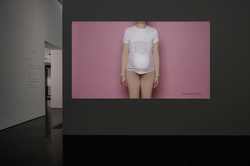 a projected video shows the torso of a pregnant person who is wearing only a stretched white t-shirt and stands in front of a pink backdrop