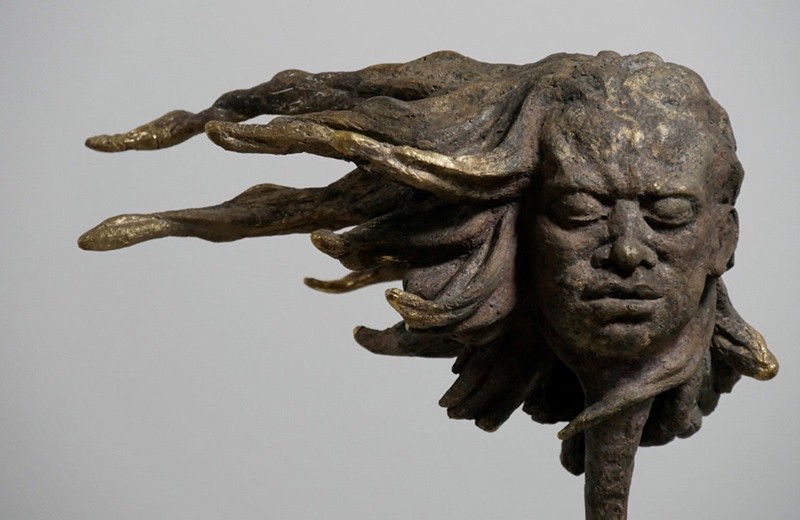 A bronze sculpture of a human head with its eyes closed and hair standing straight out to the left side.