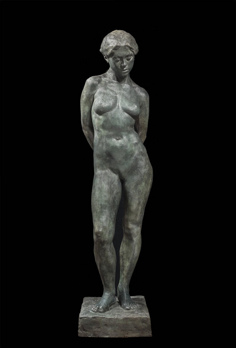 A bronze sculpture of a female figure standing with her arms held behind her back. Her gaze is directed toward the floor.