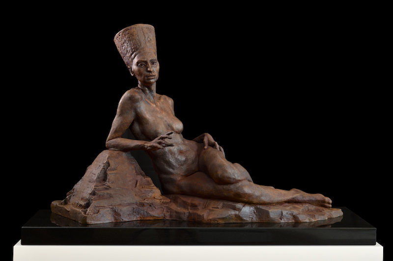 A bronze sculpture of a female pharaoh reclining against what seems to be a miniature pyramid. The sculpture is placed atop a black pedestal.