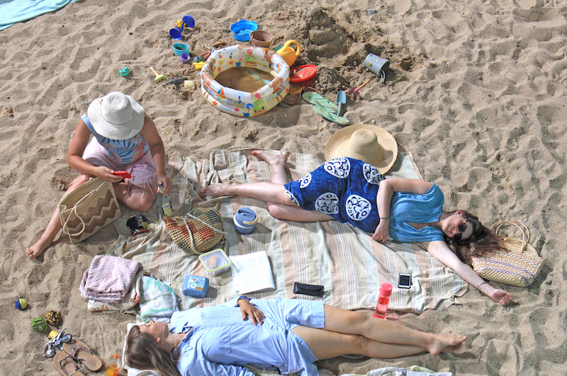 several people lie on a sandy beach, on towels, with beach toys spread around