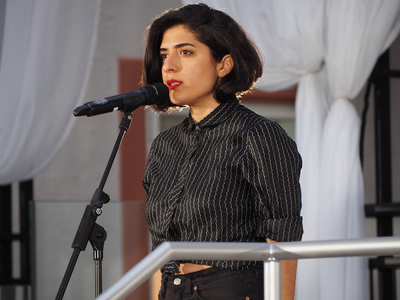 tanasgol sabbagh, a person with dark bobbed hair and red lipstick, speaks into a microphone on stage