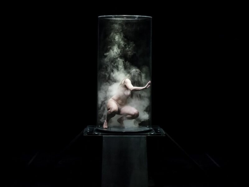 A photo of a naked woman in immersed in water a transparent tank placed in a black room.