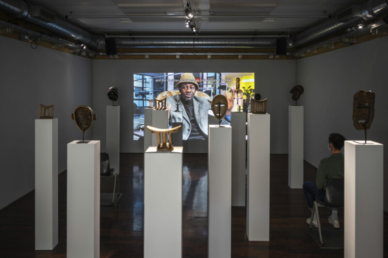 Exhibition view showing a sculpture installation of masks on plinths and a video screened on the wall behind it