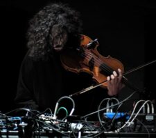 a person with dark curly hair leans over a desk with wires and cables, attached to a violin in his hand