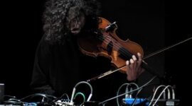 a person with dark curly hair leans over a desk with wires and cables, attached to a violin in his hand