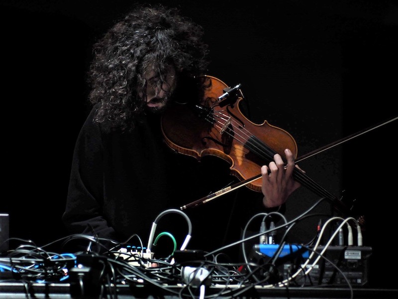 a person with dark curly hair leans over a desk with wires and cables, attached to a violin in his hand