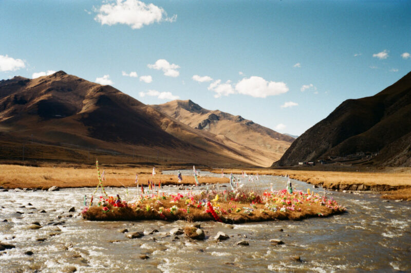 A photo of a Tibetan landscape with hills, a stream and a patch of land in the middle with colorful ceremonial decorations