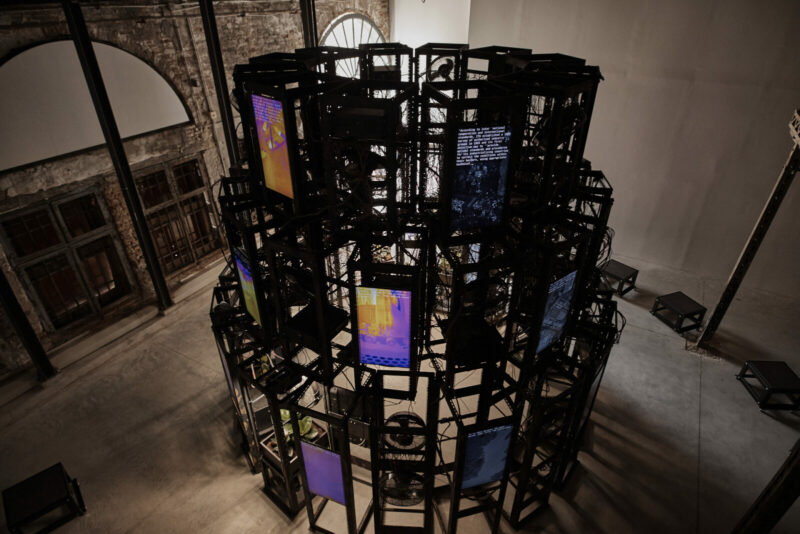 A photo of an art installation - a tall, black cylindrical construction holding several screens