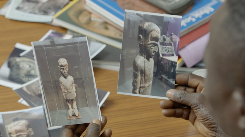 two hands hold printed images of the Balot statue