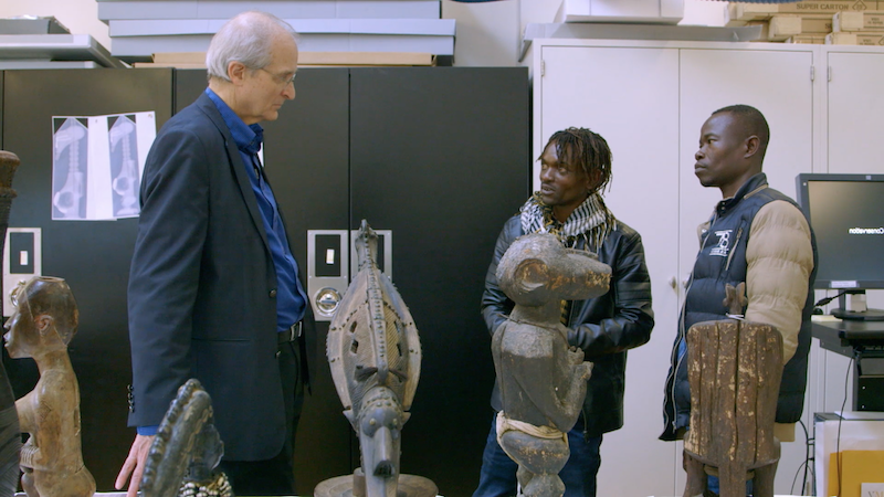 two members of CATPC stand in a university classroom amid African busts, speaking to a tall white man with white hair