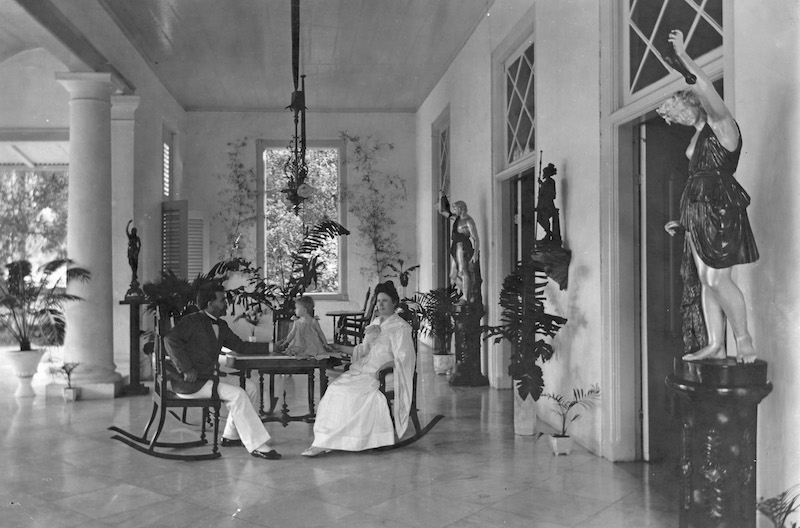 a black and white photo of a man and woman all dressed in white sitting on a luxurious veranda