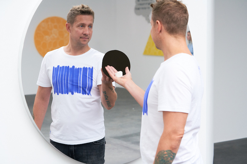 Jeppe Hein stands in front of his mirrored installation with his hands in front of a hole, holding a small piece of chalk