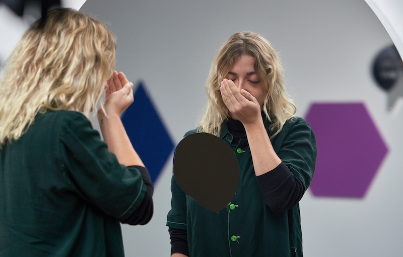 a blond haired woman stands in front of the mirrored installation, sniffing her palm with her eyes closed
