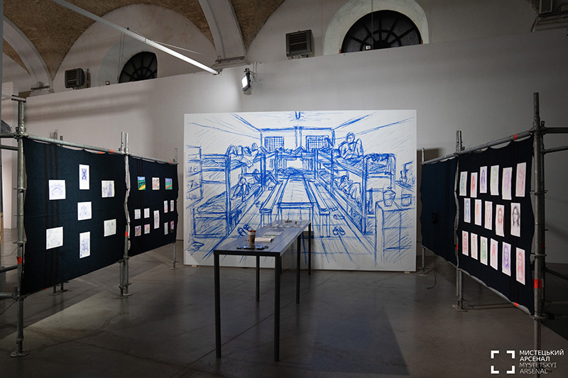 A long table in front of a white wall upon which a large blue drawing has been made.