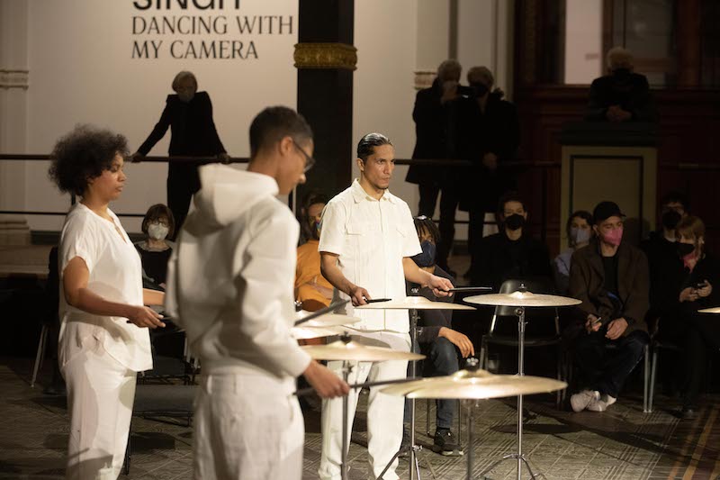 a group of cymbal players, all dressed in white, stand around the atrium of Gropius Bau playing their cymbals
