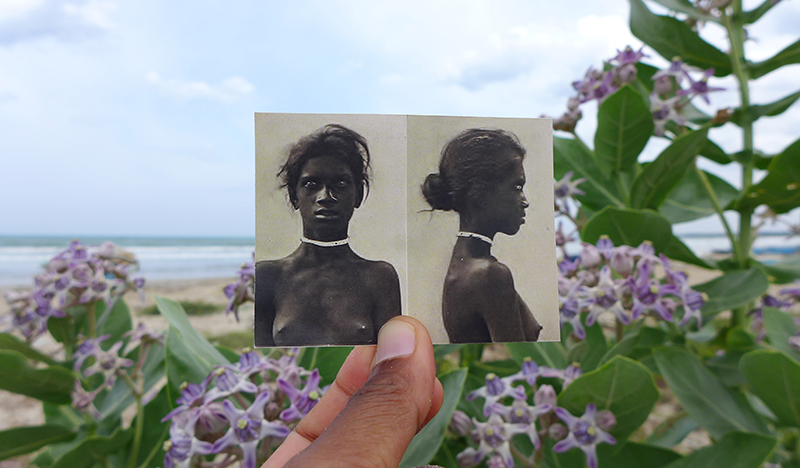 A hand holding a black and white "mug-shot" like photograph of an indigenous Sri Lanken woman with a beautiful beach scene in the background