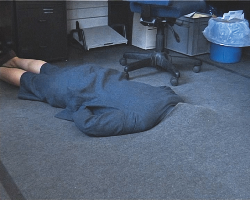 A female person dressed in grey, lying on a grey carpet that seems to swallow her head