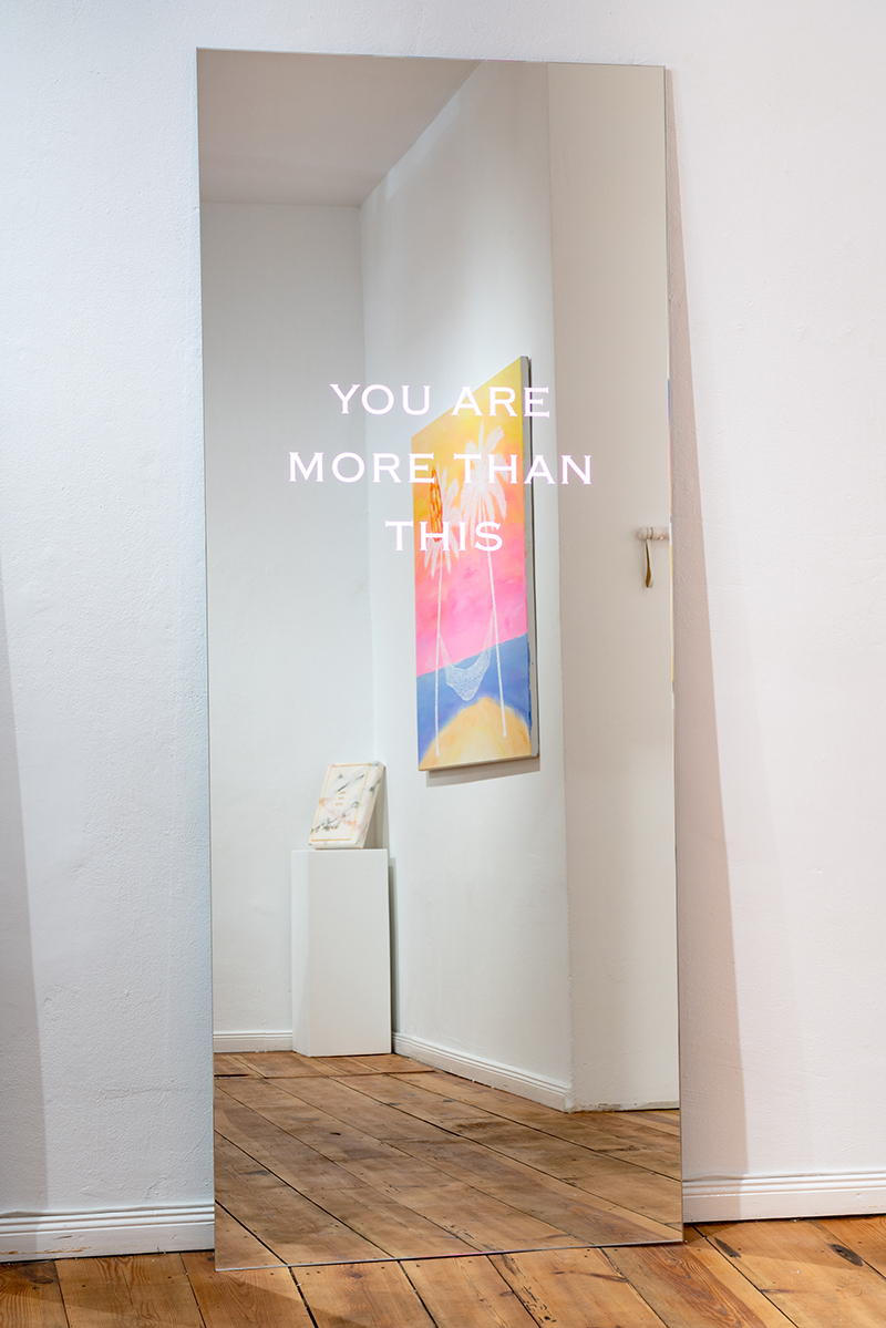 A photograph of a floor-length mirror leaning against the wall with the words "you are more than this" on it