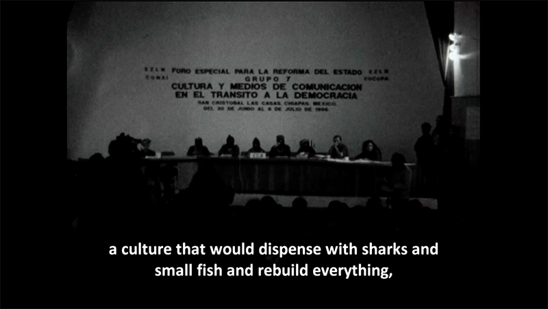 A video still of masked men sitting on stage at a conference table with subtitles that read, "a culture that would dispense with sharks and small fish and rebuild everything."