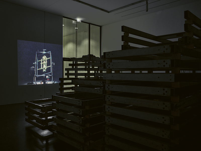 Photo of a sculpture made from stacked aluminium frames. A film is being projected behind. The room is dark.