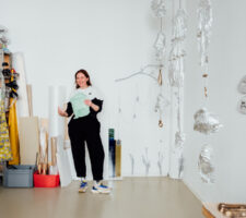 Artist Vanessa Brown stands laughing in a white studio with tin foil sculptures hanging from the ceiling