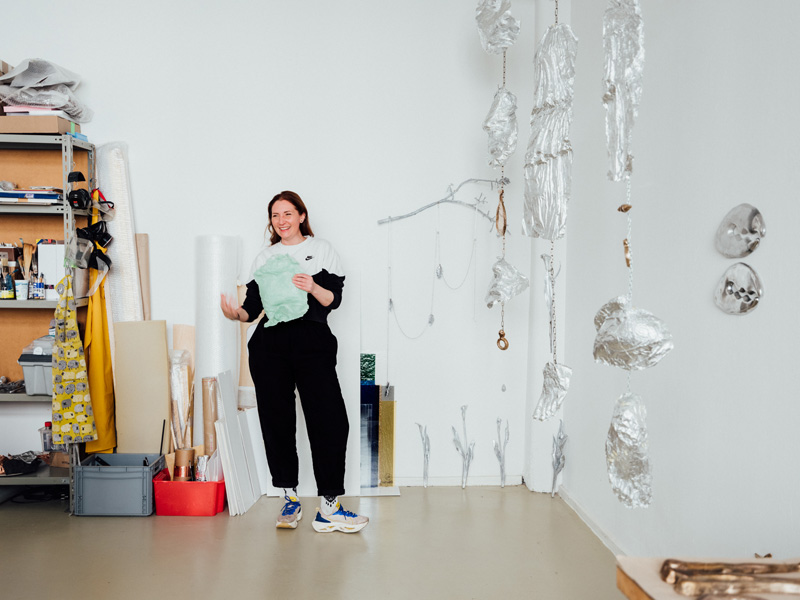 Female artist stands laughing in a white studio with tin foil sculptures hanging from the ceiling