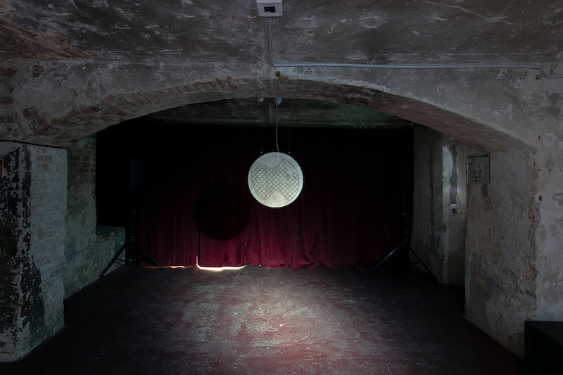 dark room with concrete walls and floor, a red curtain hangs over the fall wall and a white circular disc hangs in front