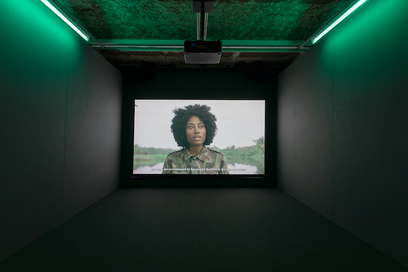 a dark cinematic room with a large screen to one end, where a woman dressed in army green is speaking to the camera