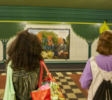 two women stood on the platform of an underground station looking at an artwork on the wall